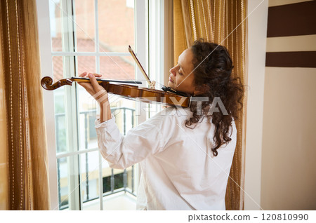Woman playing violin by window with peaceful expression indoors 120810990