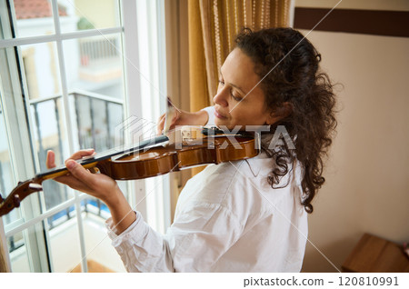 Woman playing violin near window with peaceful expression and natural light 120810991