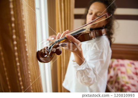 Woman playing violin by window in warm, softly lit room 120810993