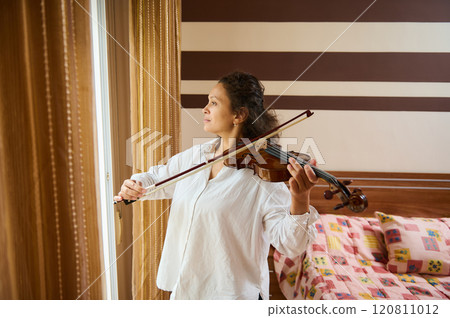 Woman playing violin by a window in a cozy room setting Woman playing violin by a window in a cozy room setting 120811012
