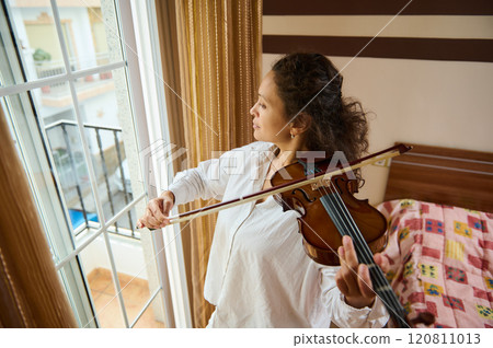 Woman playing violin near window in a cozy home environment Woman playing violin near window in a cozy home environment 120811013