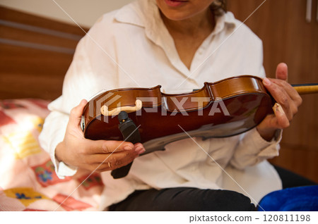 Woman tuning a violin in a cozy bedroom setting during daylight Woman tuning a violin in a cozy bedroom setting during daylight 120811198