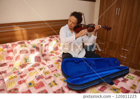 Woman playing violin while sitting on bed in cozy bedroom setting Woman playing violin while sitting on bed in cozy bedroom setting 120811225