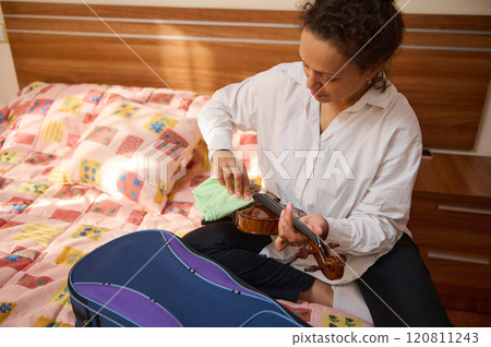 Woman sitting on bed polishing a violin with a cloth in hand Woman sitting on bed polishing a violin with a cloth in hand 120811243