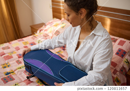 Woman packing blue suitcase on colorful bed in well-lit room Woman packing blue suitcase on colorful bed in well-lit room 120811657
