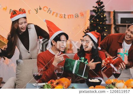 Group of young Asian man and women as friends having fun at a New Year's celebration, holding gift boxes standing by Christmas tree decoration, midnight countdown Party at home with holiday season. 120811957