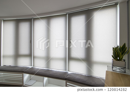 Motorized roller shades in the interior. Automatic roller blinds beige color on big glass windows. Home luxury curtains are above the windowsill with pillows. Smart house. Green trees outside. 120814282