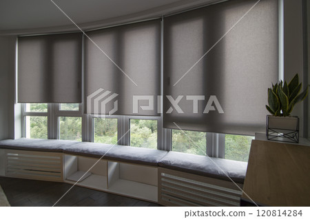 Motorized roller shades in the interior. Automatic roller blinds beige color on big glass windows. Home luxury curtains are above the windowsill with pillows. Smart house. Green trees outside. Motorized roller shades in the interior. Automatic roller blinds beige color on big glass windows. Home luxury curtains are above the windowsill with pillows. Smart house. Green trees outside. 120814284