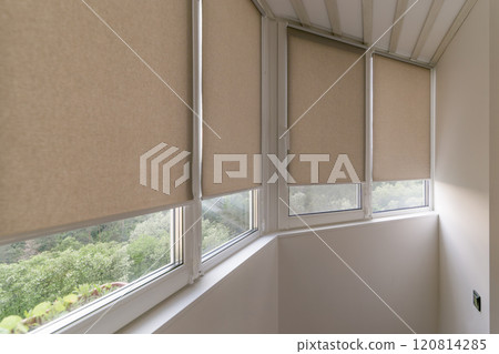Modern roller blinds in a neutral beige color covering windows in a bright enclosed balcony space. Natural light filters through roller shades, with a greenery outside, creating a cozy atmosphere. Modern roller blinds in a neutral beige color covering windows in a bright enclosed balcony space. Natural light filters through roller shades, with a greenery outside, creating a cozy atmosphere. 120814285