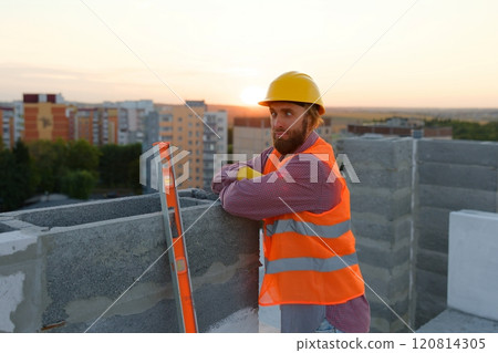 Construction worker wearing hardhat and safety vest leaning on concrete block at sunset Construction worker wearing hardhat and safety vest leaning on concrete block at sunset 120814305