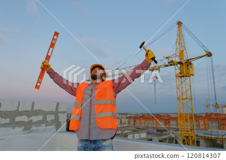 Construction worker celebrating with level and hammer at building site Construction worker celebrating with level and hammer at building site 120814307