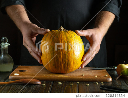Hands gently cradle a bright orange pumpkin, ready for cooking. The rustic wooden table adds warmth, complemented by kitchen tools and a hint of green apples, evoking autumn creativity Hands gently cradle a bright orange pumpkin, ready for cooking. The rustic wooden table adds warmth, complemented by kitchen tools and a hint of green apples, evoking autumn creativity 120814834