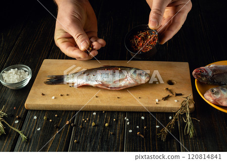 Hands expertly season a fresh fish with spices on a wooden cutting board. Surrounding ingredients include salt and herbs, creating an inviting atmosphere for culinary creativity. 120814841
