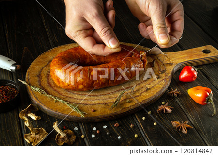 Hands expertly tie a freshly made sausage on a wooden board. Vibrant peppers and fragrant herbs surround the savory creation, showcasing culinary artistry and traditional methods Hands expertly tie a freshly made sausage on a wooden board. Vibrant peppers and fragrant herbs surround the savory creation, showcasing culinary artistry and traditional methods 120814842