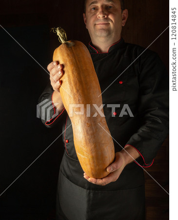 In a cozy kitchen, a confident chef proudly holds a giant butternut squash, highlighting the beauty of seasonal produce. The warm light enhances the earthy tones of the vegetable 120814845