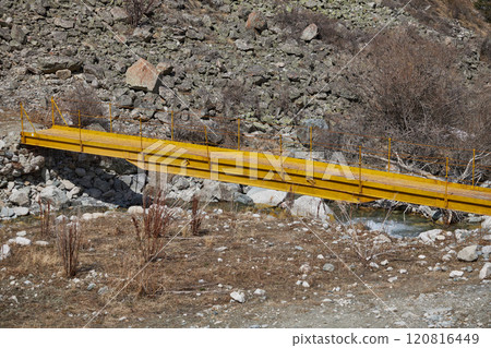 A yellow metal pedestrian bridge connects the banks of a small river. A yellow metal pedestrian bridge connects the banks of a small river. 120816449