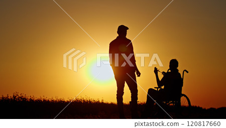 Silhouette of a person standing near a woman in a wheelchair at sunset. A symbolic image of support, care, and companionship in a serene rural setting with a warm golden glow. 120817660