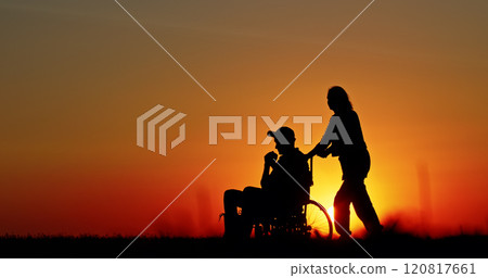Silhouette of a person pushing a wheelchair during a vibrant sunset, symbolizing care, assistance, and support in a tranquil rural setting. The golden sky enhances the emotional warmth. 120817661