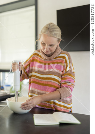 A focused middle-aged woman in a colorful striped sweater uses a hand blender to mix ingredients in a white bowl, preparing a recipe in her kitchen 120817728