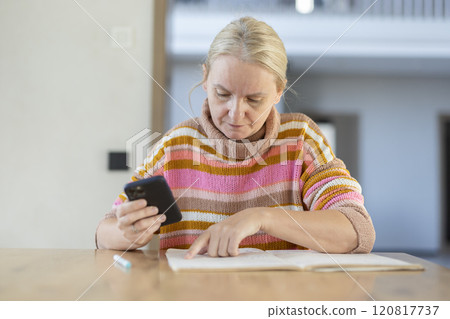 A middle-aged woman in a colorful striped sweater studies a book while holding a smartphone, sitting at a wooden table in a well-lit room. 120817737