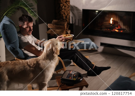 A young man relaxes by the fireplace with a plate of waffles, while a Golden Retriever stands nearby, creating a warm and cozy atmosphere. A young man relaxes by the fireplace with a plate of waffles, while a Golden Retriever stands nearby, creating a warm and cozy atmosphere. 120817754