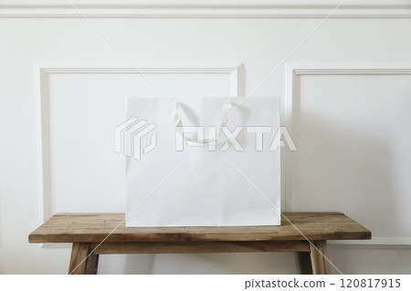 Clean simple white paper shopping bag mockup with handles on old wooden table, bench. White wall background with stucco decor. Empty gift bag for branding display, message. Retail business concept 120817915