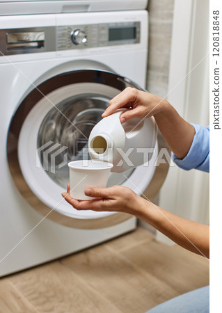 Woman pours liquid transparent laundry detergent or conditioner into plastic cap against washing machine. 120818848