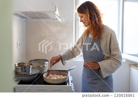 A woman is frying pieces of chicken breast in a pan on an induction stove, stirring them with a silicone spatula. 120818861