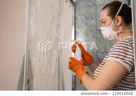 A woman is actively cleaning her shower using a spray bottle and wearing protective gloves 120818888