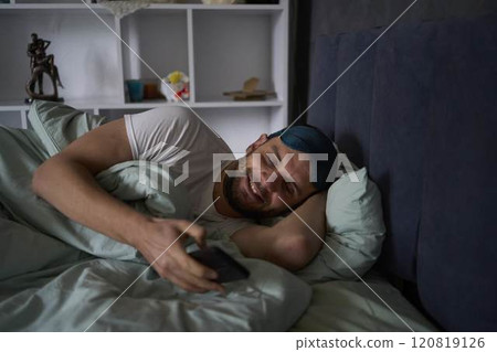30-year-old man wearing a silk sleep mask is chatting on a smartphone and smiling 120819126