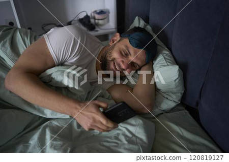 30-year-old man wearing a silk sleep mask is chatting on a smartphone and smiling 30-year-old man wearing a silk sleep mask is chatting on a smartphone and smiling 120819127