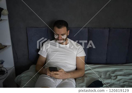A 30-year-old man with moisturizing patches under his eyes is chatting on a smartphone 120819129