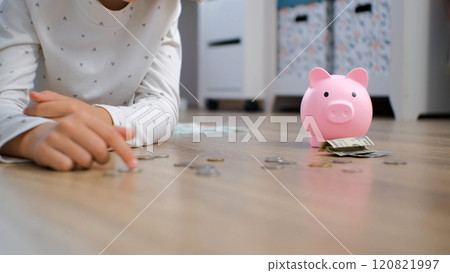 Close-up of young girl lying on the floor of her cozy room sorting coins with a pink piggy bank, enjoying her first steps in saving Close-up of young girl lying on the floor of her cozy room sorting coins with a pink piggy bank, enjoying her first steps in saving 120821997