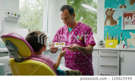 A cheerful male dentist in his 40s, wearing a colorful scrub, interacting with a young child patient (around 8 years old) in a fun, colorful dental office. The dentist is offering a treat to the child A cheerful male dentist in his 40s, wearing a colorful scrub, interacting with a young child patient (around 8 years old) in a fun, colorful dental office. The dentist is offering a treat to the child 120823549