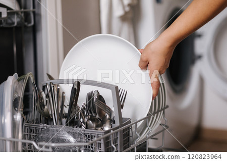 Side view of human hand removing clean plates from a loaded dishwasher. Background of open wash machine  120823964