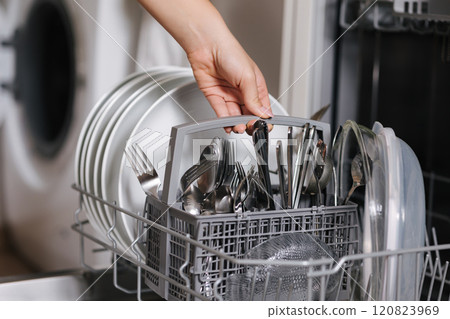 Female hand removing clean cutlery from a loaded dishwasher. Background of open wash machine  120823969