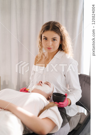 Skincare specialist distributes the clay mask thoroughly over the client's face, ensuring even coverage for a balanced complexion Skincare specialist distributes the clay mask thoroughly over the client's face, ensuring even coverage for a balanced complexion 120824004
