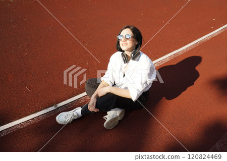 Young woman in casual outfit and headphones sits on a track in the sunlight, enjoying music and relaxation outdoors. 120824469