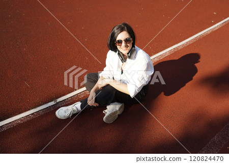 Young woman in casual outfit and headphones sits on a track in the sunlight, enjoying music and relaxation outdoors. 120824470