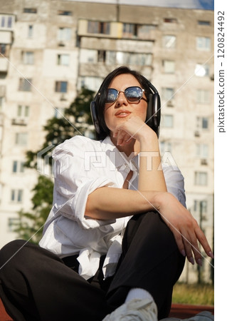 Woman in sunglasses and headphones sits outdoors on a track, enjoying a sunny day and her favorite tunes, vertical photo. 120824492