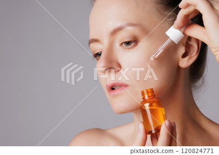 Happy smiling woman applying serum on her face, close eyes with pleasure. Redhead girl holding dropper with skin care product for healthy and glowing skin tone Happy smiling woman applying serum on her face, close eyes with pleasure. Redhead girl holding dropper with skin care product for healthy and glowing skin tone 120824771