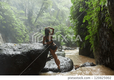 A young slender woman in a black swimsuit poses on a large rock in the water in the rain near a jungle waterfall on the popular island of Bali. A young slender woman in a black swimsuit poses on a large rock in the water in the rain near a jungle waterfall on the popular island of Bali. 120825002
