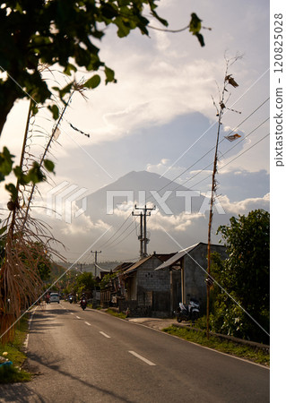 View of the volcano from the road decorated for the holiday in the village. Bamboo decorations along the road. A panorama of Mount Agung covered in clouds before sunset. View of the volcano from the road decorated for the holiday in the village. Bamboo decorations along the road. A panorama of Mount Agung covered in clouds before sunset. 120825028