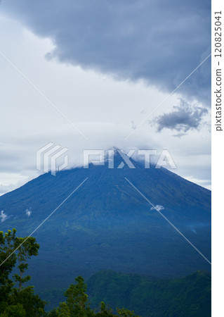 Panorama of Mount Agung and rice fields on the island of Bali. View of the mountain against a background of palm trees and a cornfield. Panorama of Agung volcano covered with clouds on a rainy day. 120825041
