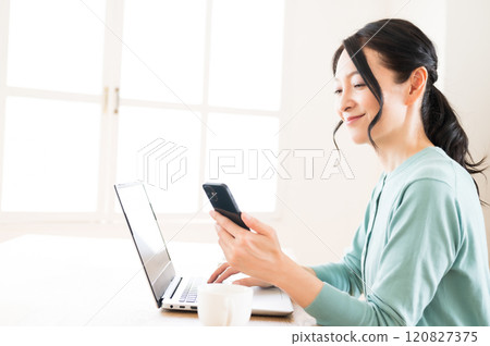 A middle-aged woman operating a computer with a smartphone in one hand 120827375