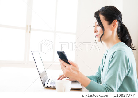 一名中年婦女單手拿著智慧型手機操作電腦 一名中年婦女單手拿著智慧型手機操作電腦 120827377
