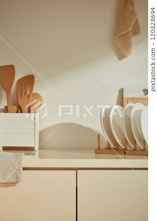 Front view photograph in minimalist kitchen. Wooden dishes and cooking utensils are featured on the beige kitchen counter, which is placed close to the wall, vacant space in center for product placing 120828694