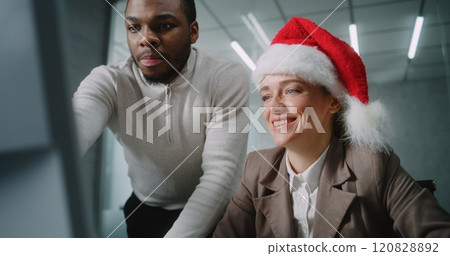 Female marketing specialist in Christmas hat uses computer with African American colleague Female marketing specialist in Christmas hat uses computer with African American colleague 120828892