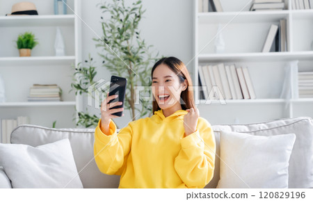 Happy young asian woman relax on comfortable couch at home texting messaging on smartphone 120829196