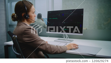 Female stock exchange trader talks on headset, monitors equity market and shares using computer 120829274